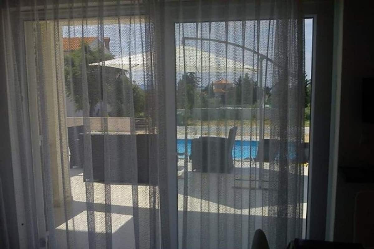 A view through a window with sheer curtains, revealing an outdoor pool area with chairs and a table.