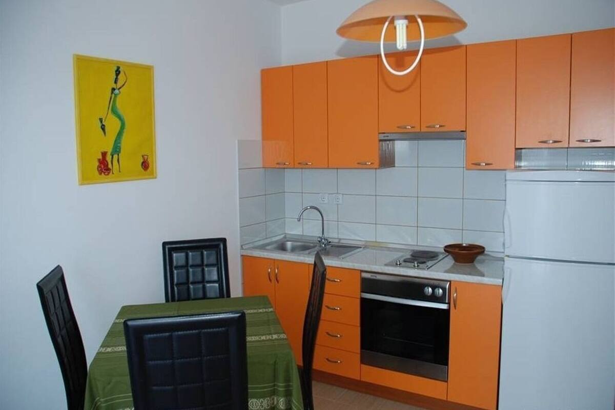 A compact kitchen with orange cabinets, a white refrigerator, a stove, and a sink. There is a dining table with chairs and a yellow artwork on the wall.