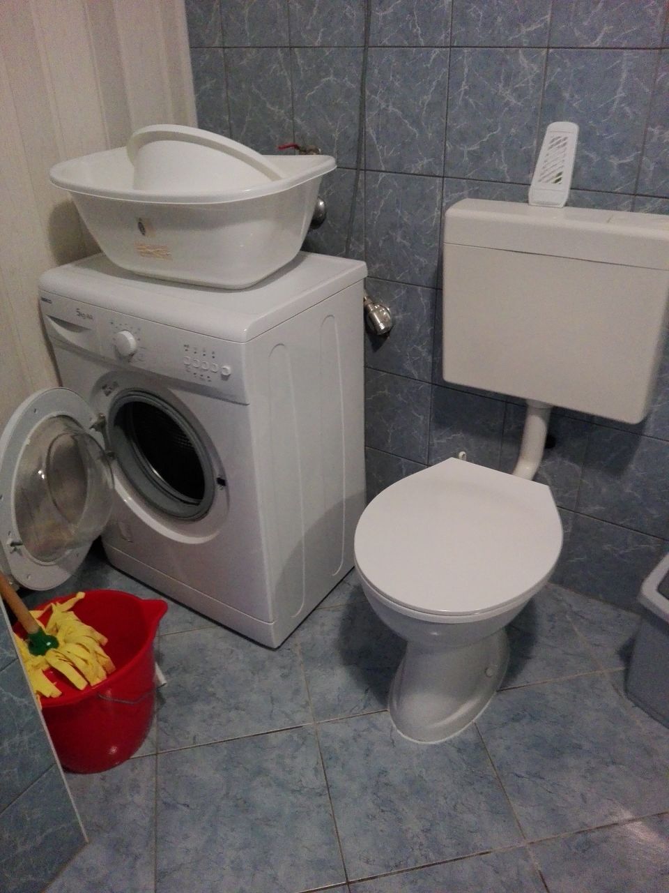 A compact bathroom with a washing machine, a toilet, and a red bucket with a green handle.