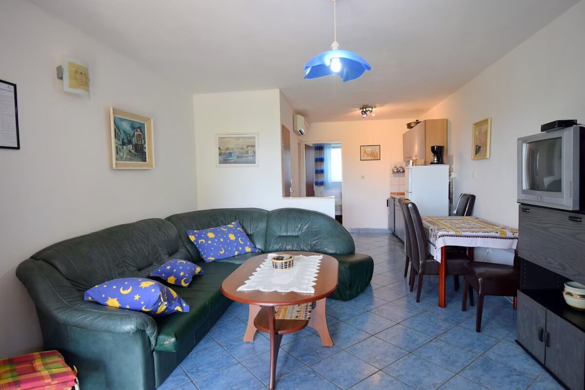 Ein Wohnzimmer mit einem grünen Sofa, einem kleinen Holztisch, einem Esstisch mit Stühlen, einem Fernseher und einer Küchenzeile im Hintergrund.
