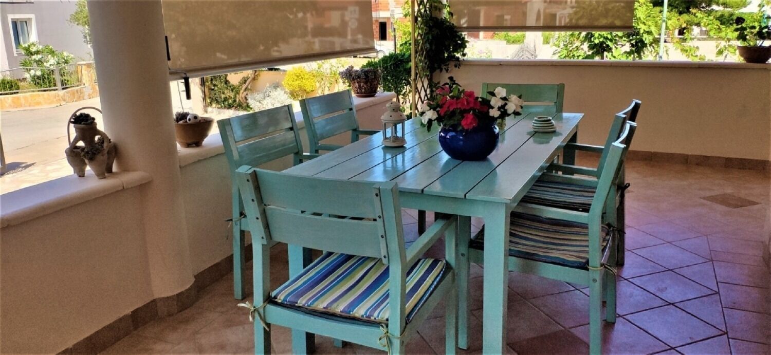 A turquoise dining table with striped cushions, surrounded by matching chairs, set on a tiled patio with potted plants and a view of the street.