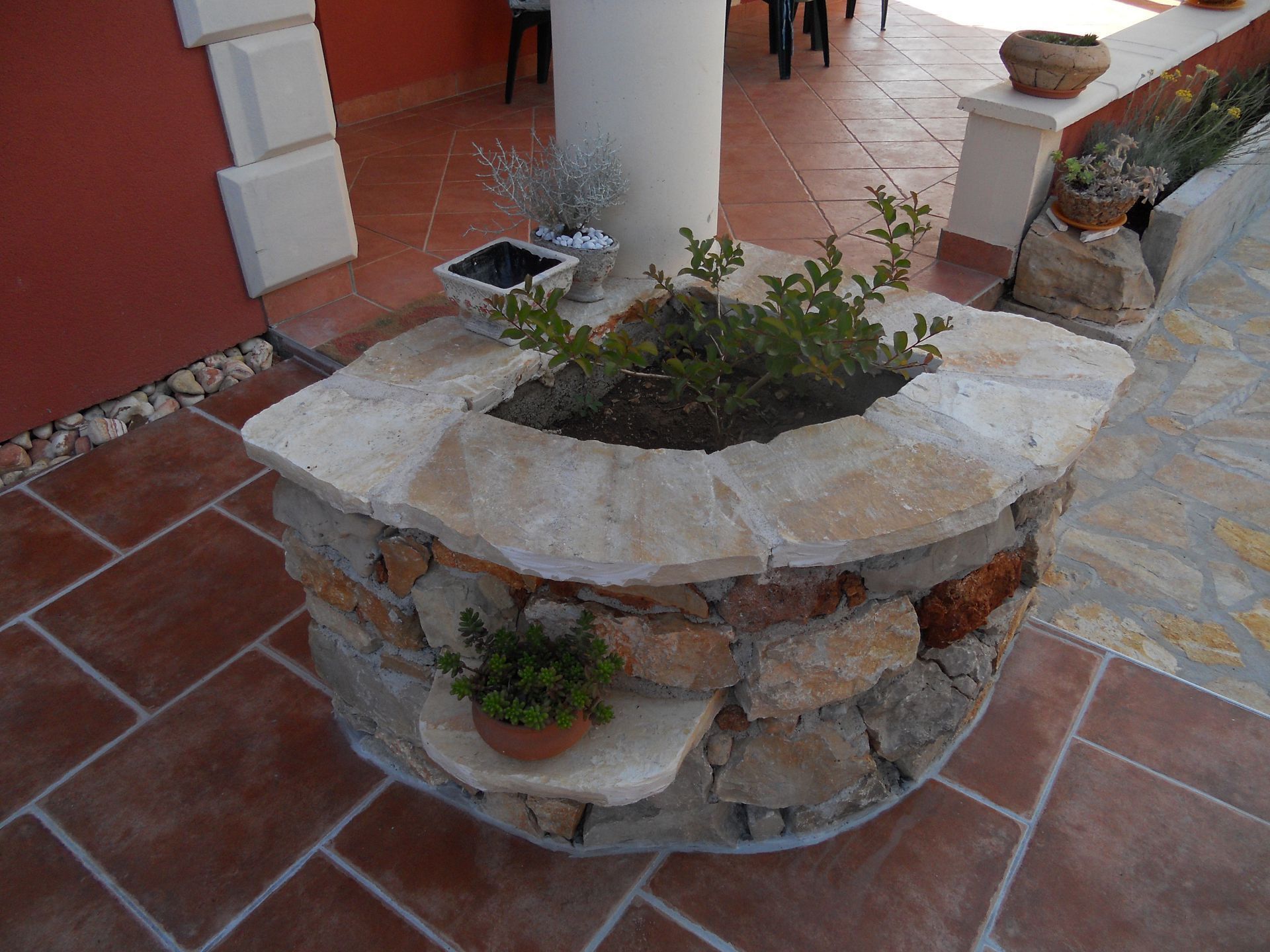 A stone planter with various plants, a small table, and a chair in the background.