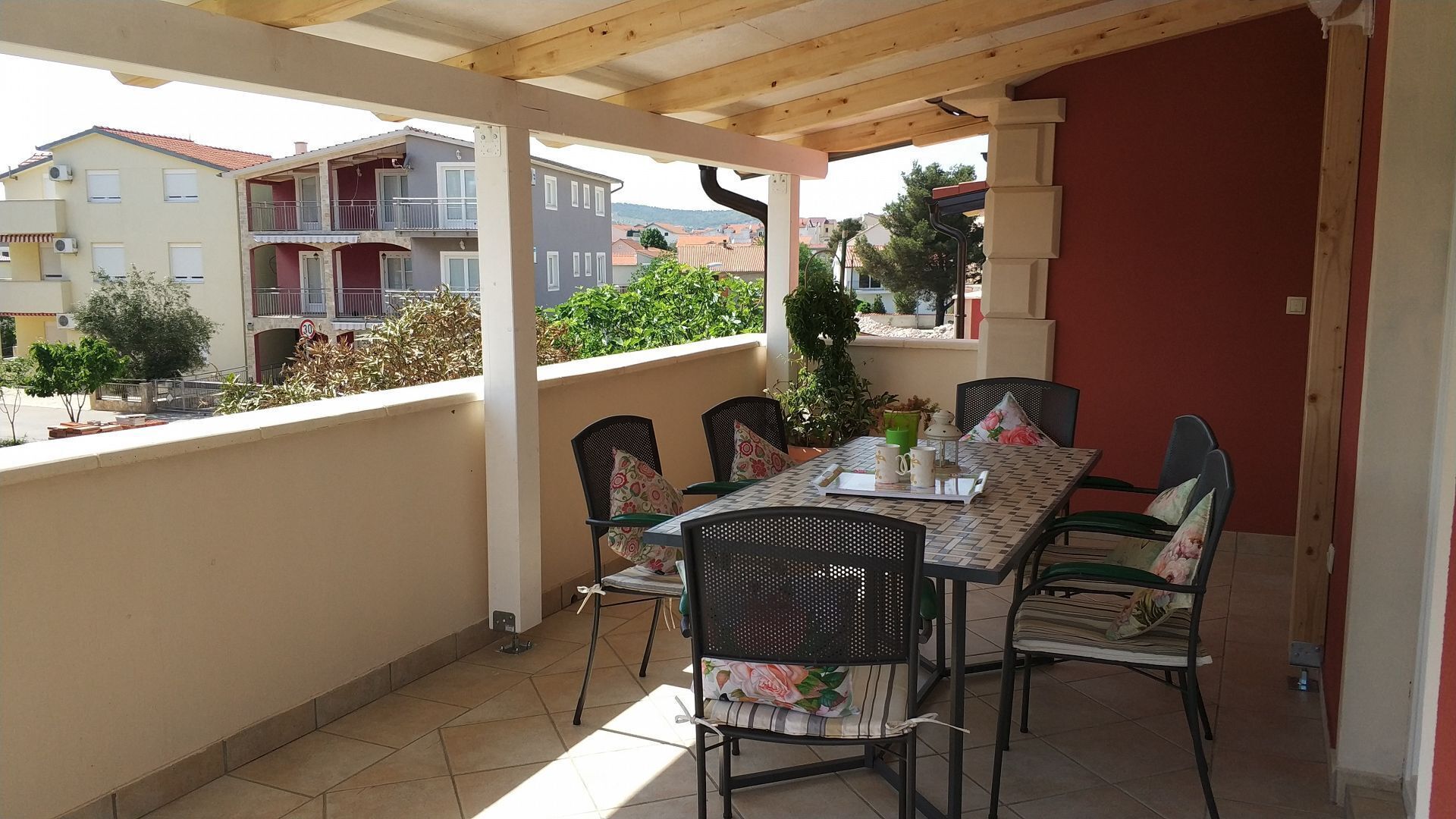 A covered outdoor dining area with a table and chairs, a view of neighboring buildings, and a red wall.