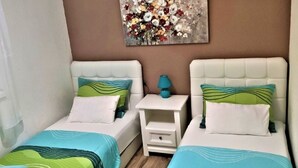 Ein Schlafzimmer mit zwei Betten, jeweils mit weißem Kopfteil und einer Bettdecke in Grün- und Blautönen. Dazwischen steht ein Nachttisch mit einer blauen Lampe und einer kleinen Schublade. An der Wand über den Betten hängt ein farbenfrohes abstraktes Gemälde.