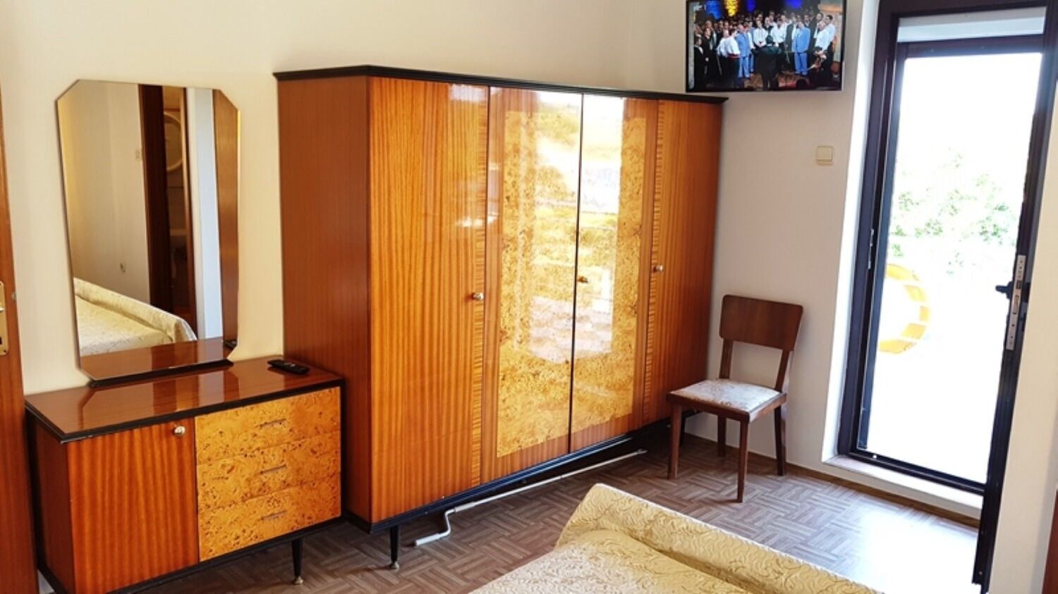 A room with a wooden wardrobe, a mirror, a chair, and a television on the wall.