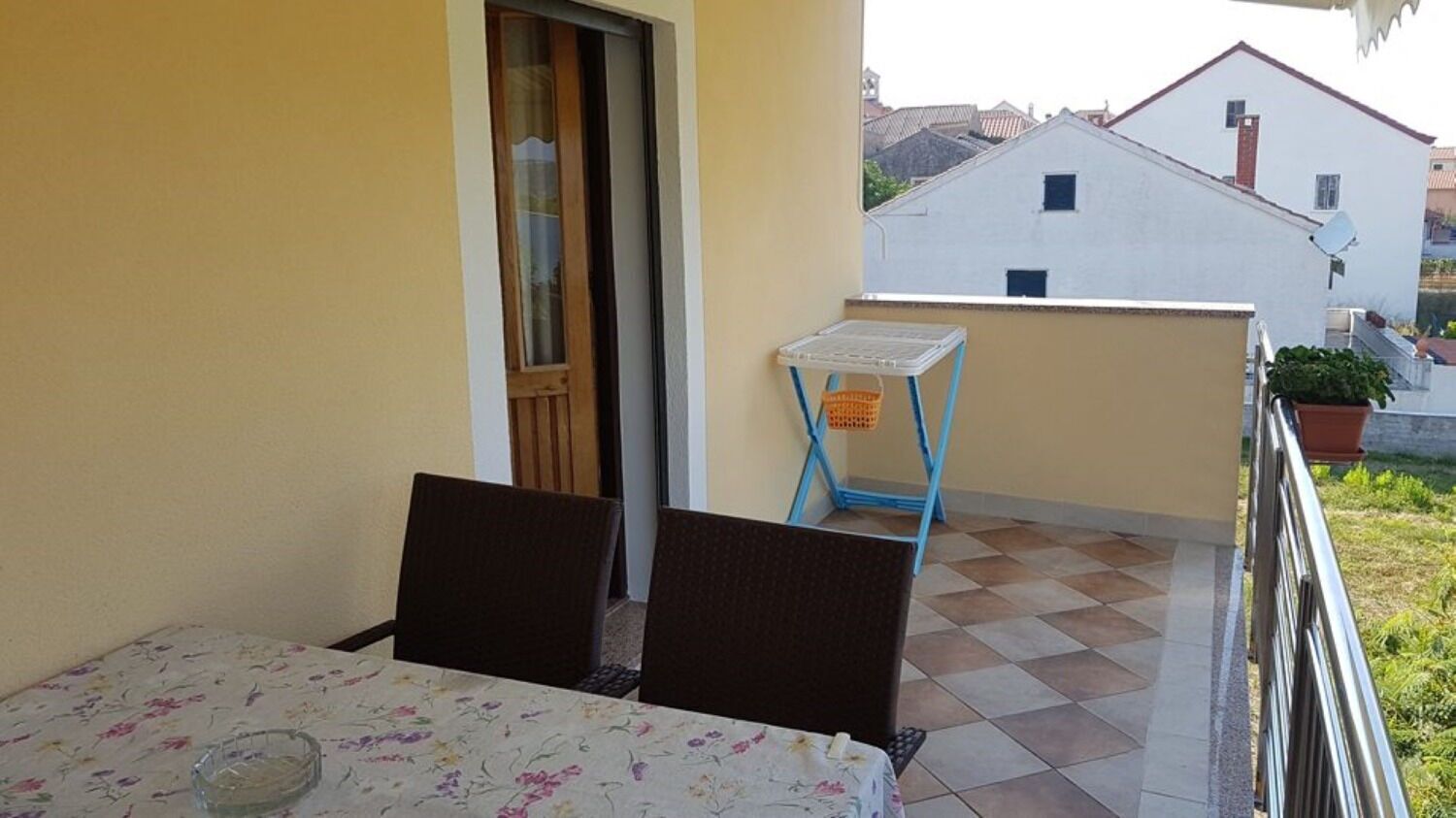 A balcony with a table and chairs, a small table with a basket, and a view of neighboring buildings.