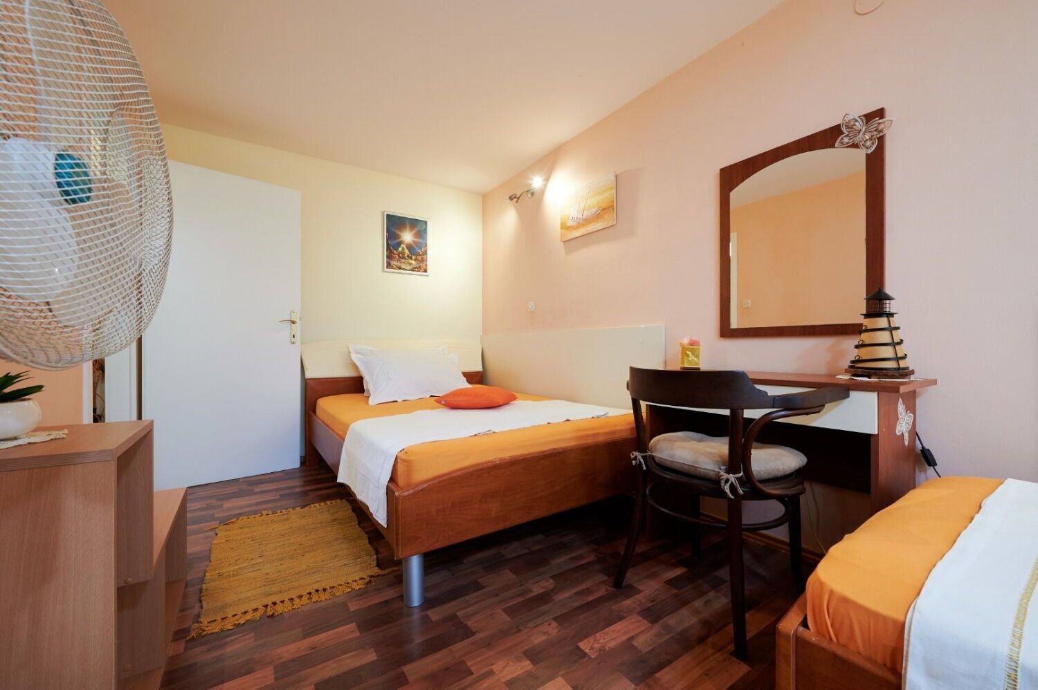 A compact hotel room with two single beds, a wooden desk, a chair, a large mirror, and a hanging lamp.