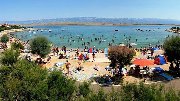 Una playa concurrida con gente nadando y tomando el sol, tiendas de campaña y barcos en el agua.