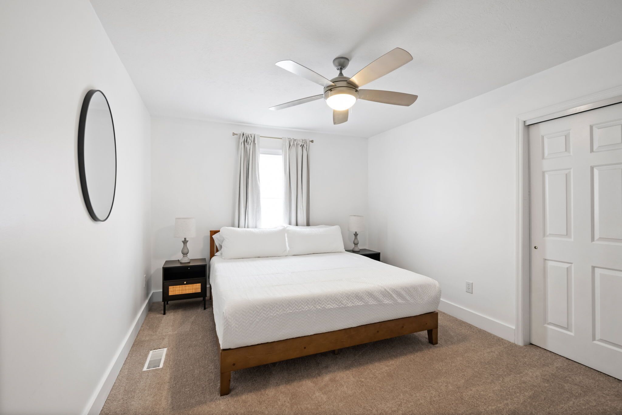 Une chambre à coucher avec un lit, un ventilateur de plafond, une table de chevet, une lampe et une fenêtre avec des rideaux.