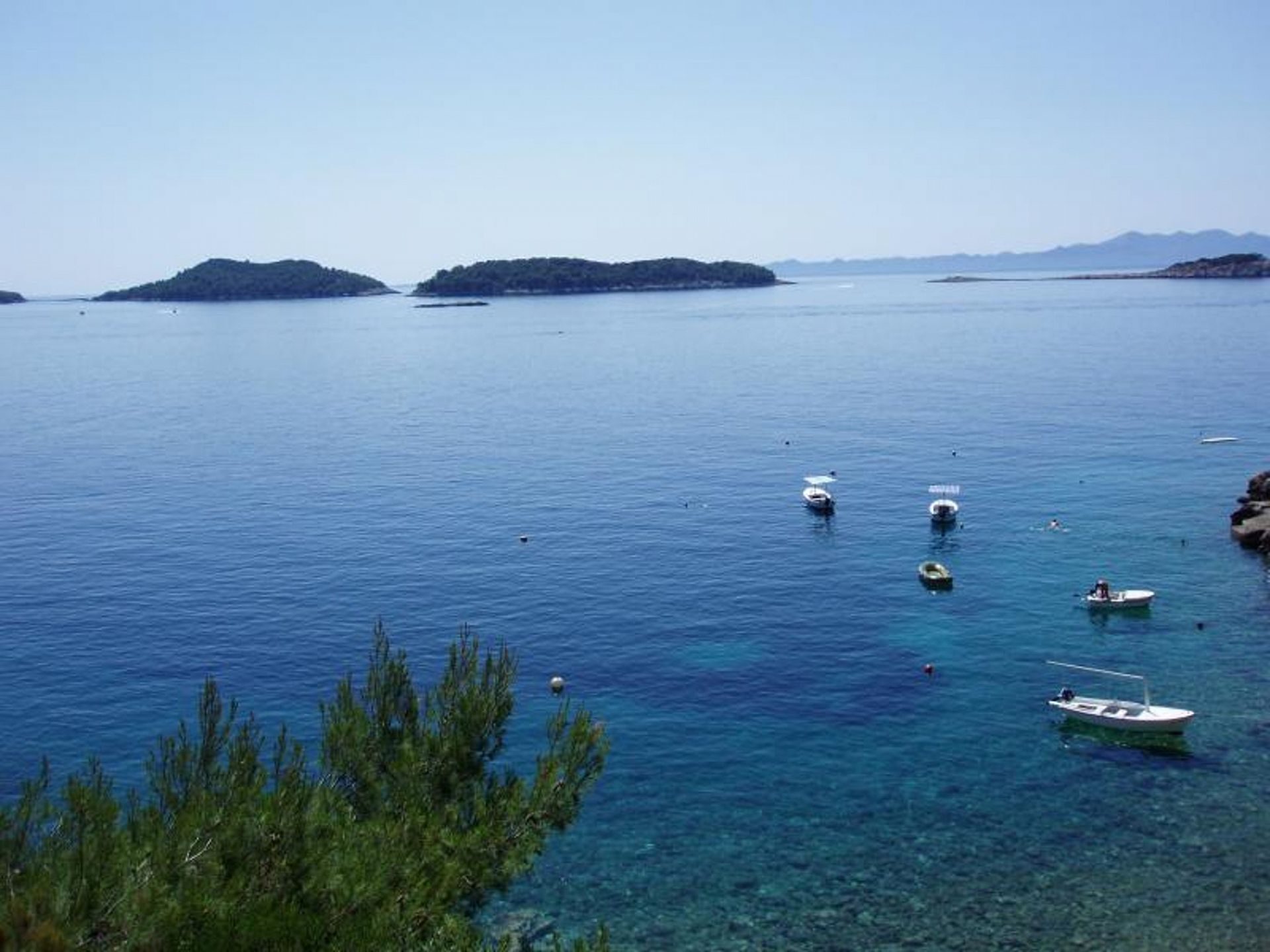 A calm sea with several small boats anchored near the shore, surrounded by clear water and distant islands.
