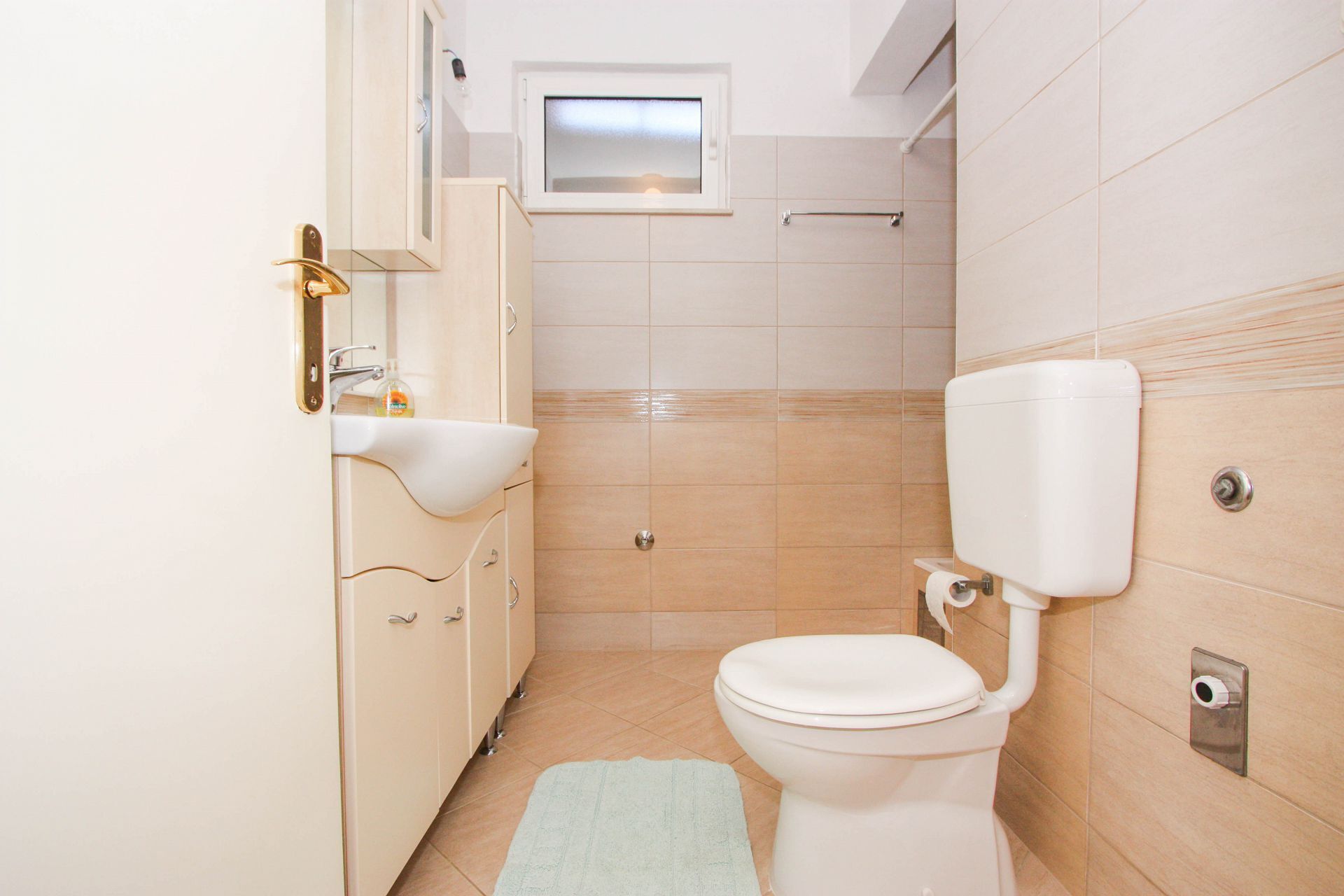 Une petite salle de bain avec des toilettes blanches, un lavabo avec un miroir et une petite fenêtre située au-dessus du miroir.