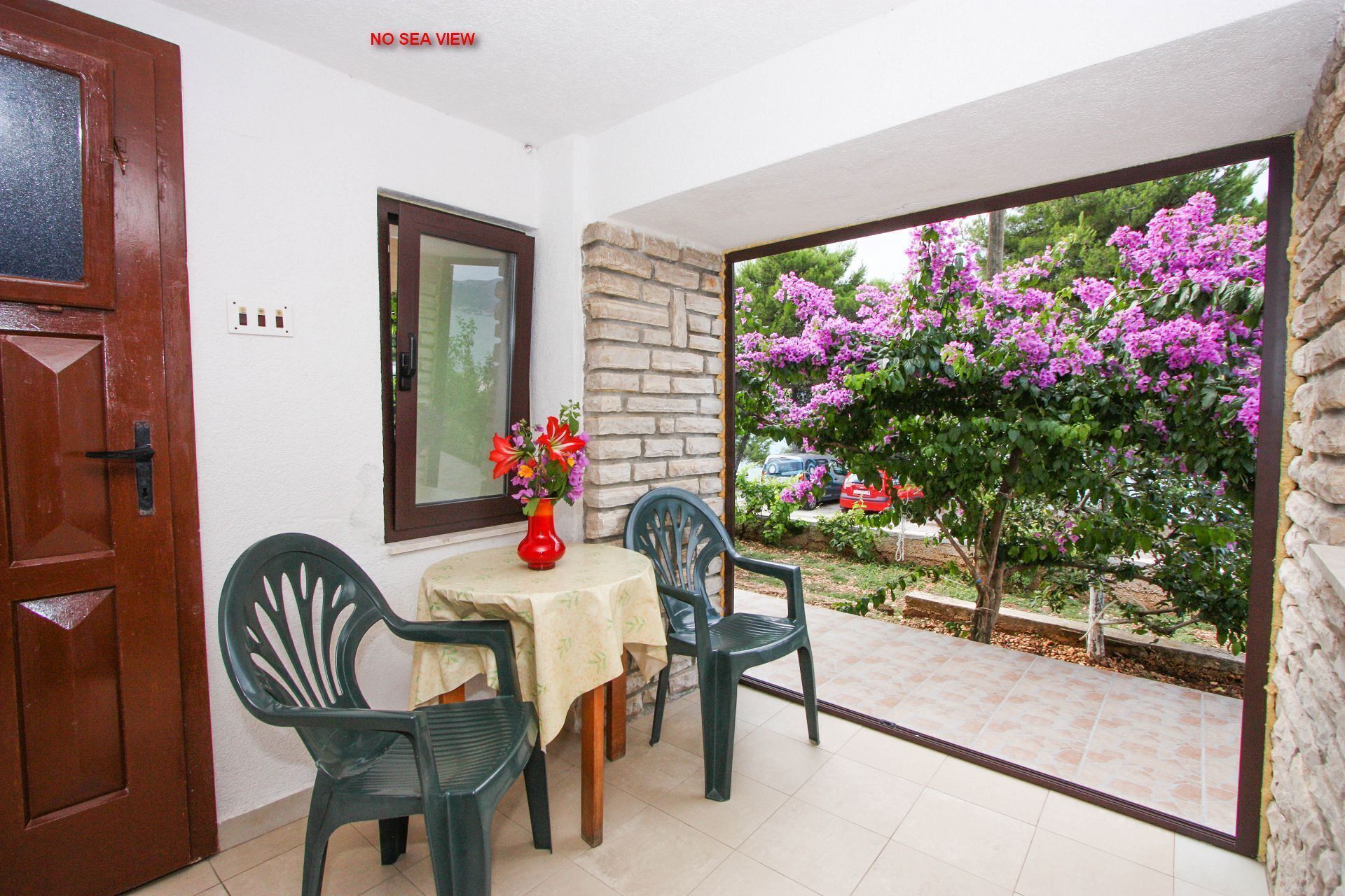 Ένα δωμάτιο με ξύλινη πόρτα, ένα παράθυρο, ένα τραπέζι με ένα βάζο λουλουδιών, δύο καρέκλες και θέα σε έναν κήπο με μωβ λουλούδια.