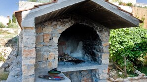 A stone-built outdoor oven with a sloped roof, surrounded by greenery and a residential building in the background. - Apartment Goran (Solta)