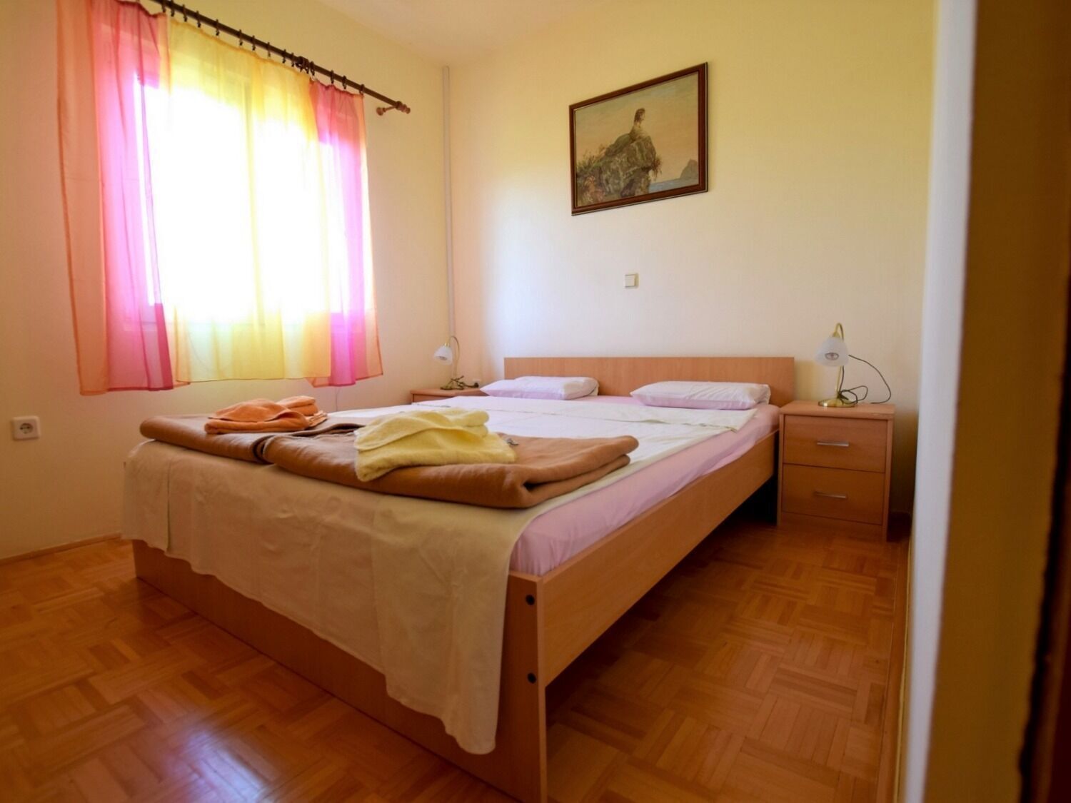 A single-bed room with a wooden floor, a bed with a white duvet and brown pillows, a nightstand with a lamp, and a painting on the wall.