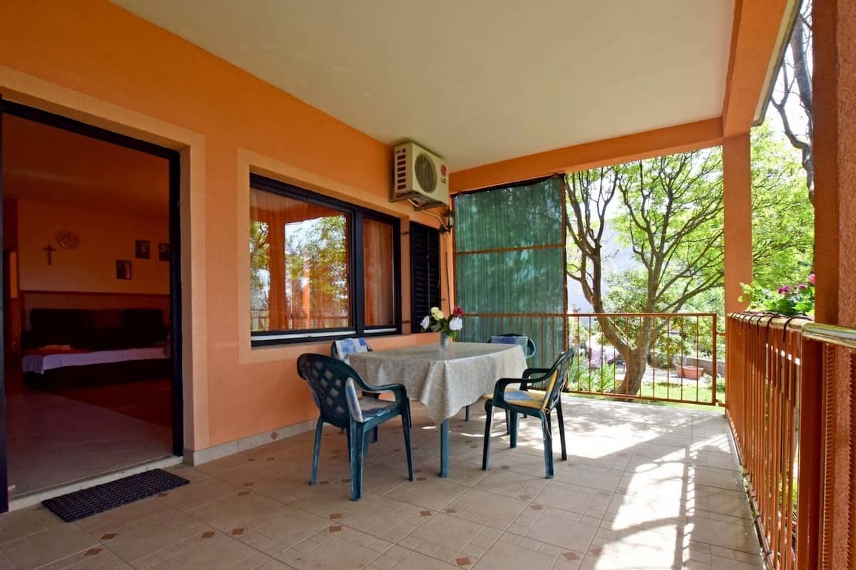 A covered patio with a table and chairs, a window with curtains, and a view of trees.