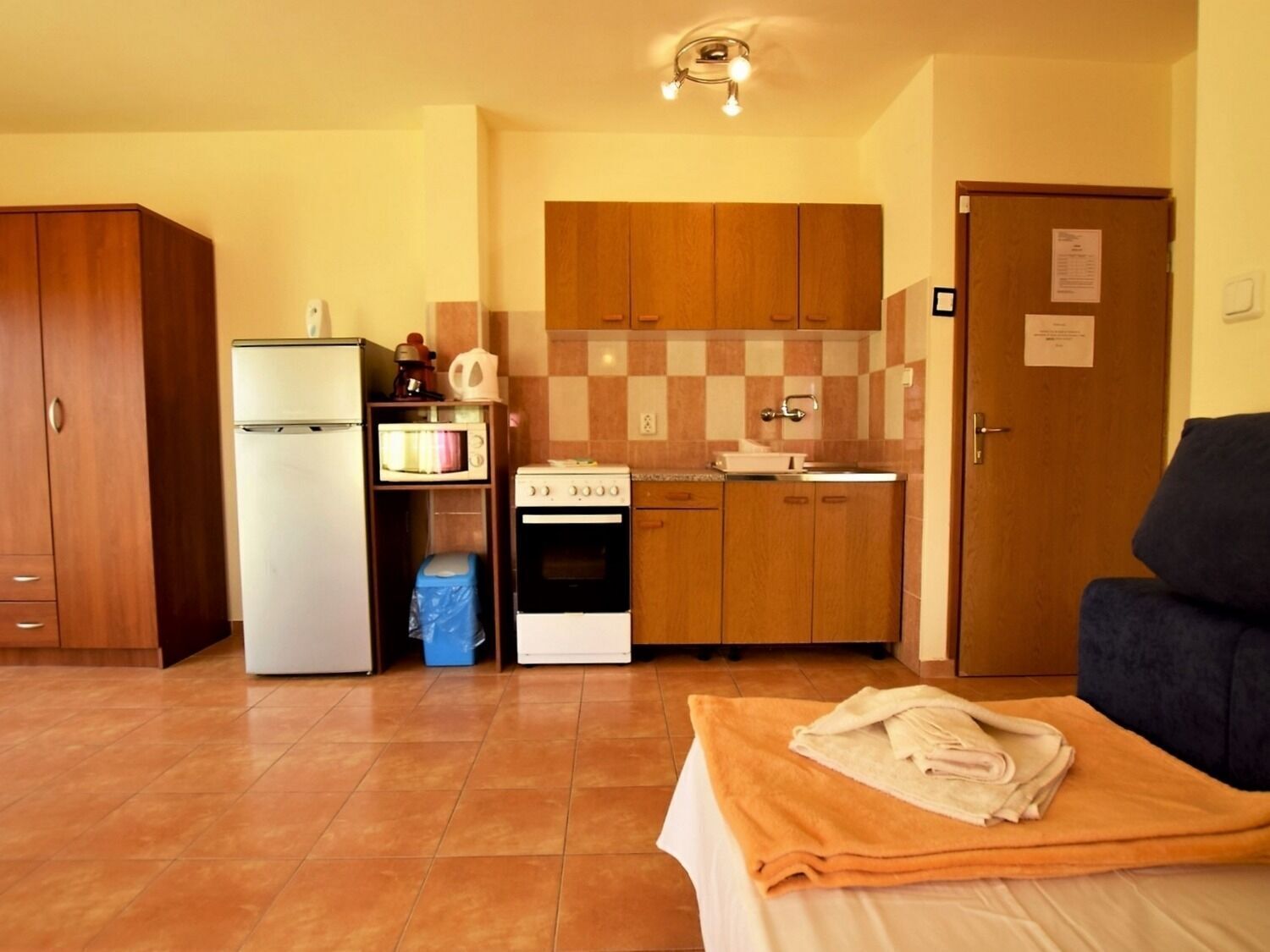 Une cuisine compacte équipée d’un réfrigérateur, d’un four à micro-ondes, d’une cuisinière et d’un évier. On y trouve un meuble en bois et une porte sur laquelle est affiché un avis.