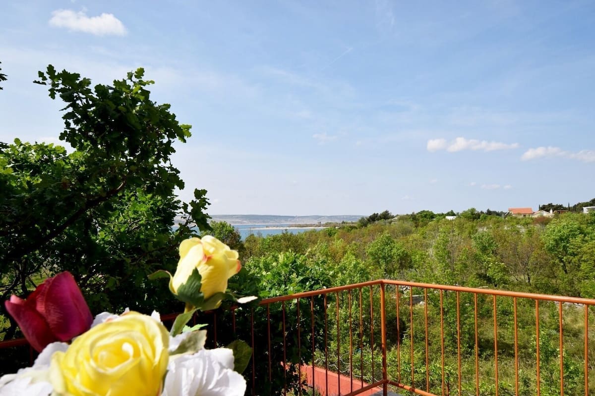 A balcony with a metal railing overlooking a lush garden and a distant view of the sea.