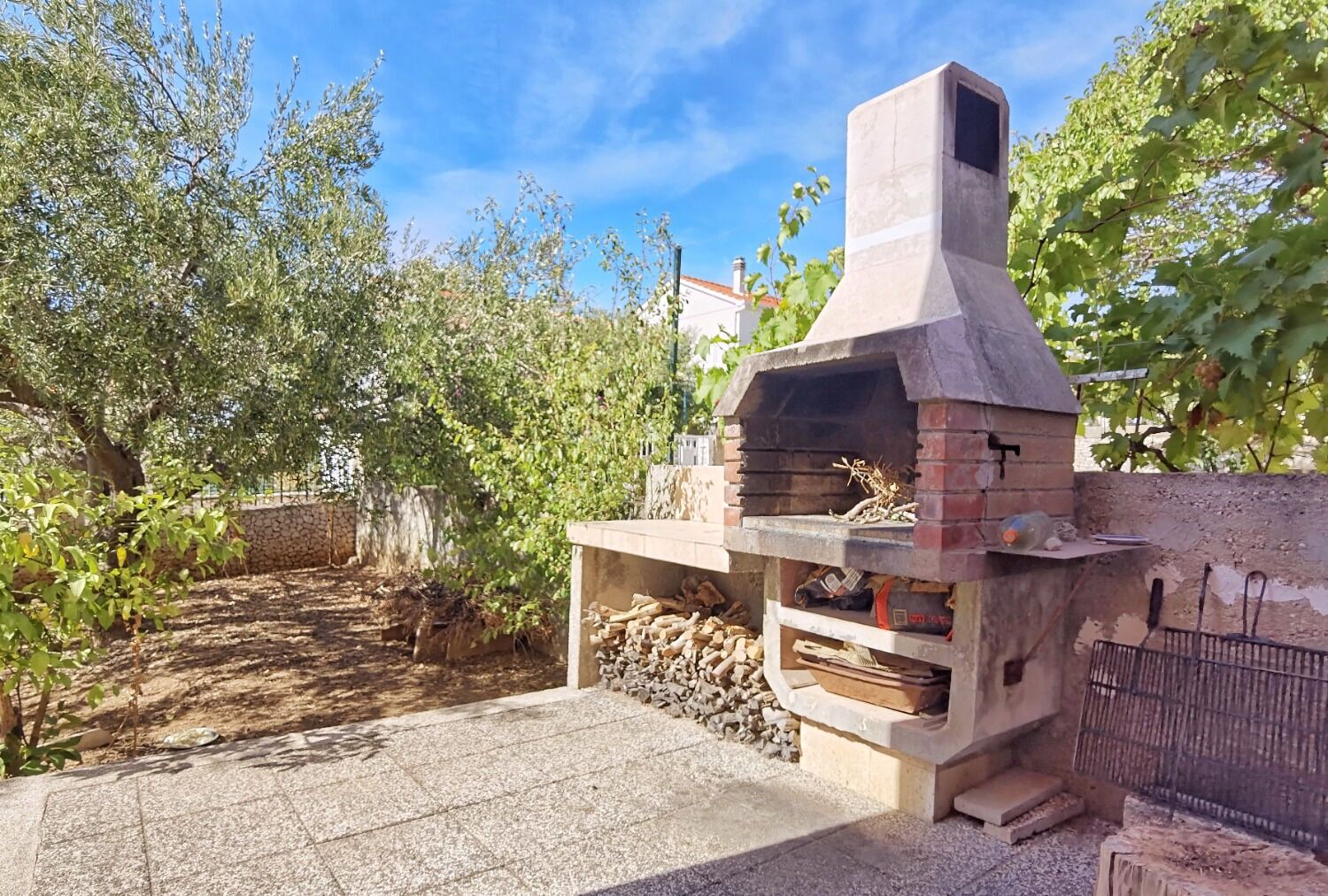 Ein Backofen aus Ziegelsteinen für Pizza, im Freien mit Schornstein, umgeben von einer Steinmauer und Grünpflanzen.