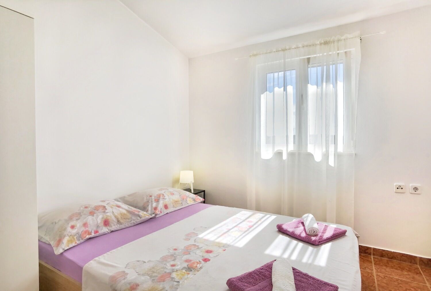 A bedroom with a bed, a nightstand, a lamp, and a window with sheer curtains.