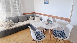 Ein modernes Wohnzimmer mit einem grauen Sofa, zwei weißen Sesseln mit gemusterten Kissen, einem kleinen runden Tisch und einem Fenster mit transparenten Vorhängen.