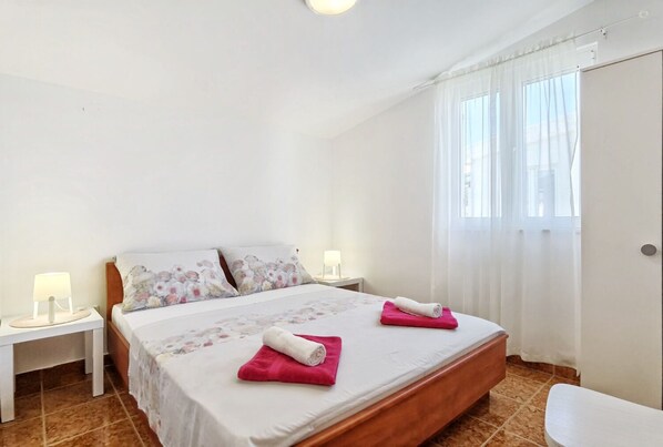A bedroom with a bed, two nightstands, a lamp, a window with curtains, and a door. - Apartments Radic (Rogoznica)