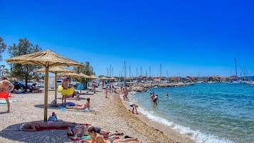 Una playa con gente tomando el sol, sombrillas y barcos al fondo.
