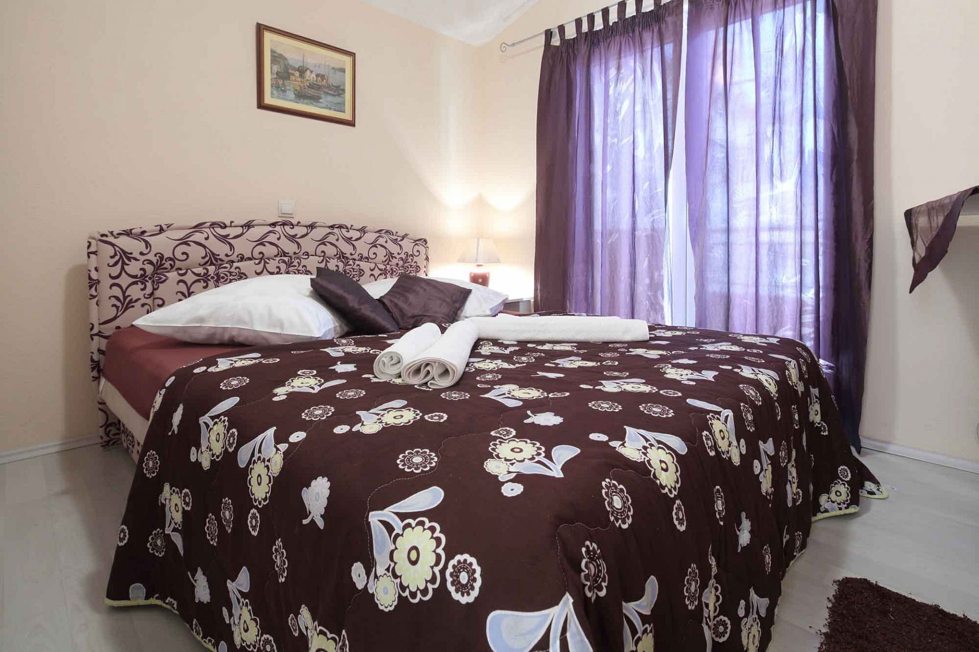 Una cama bien hecha con una colcha de estampado floral, un cabecero con diseño decorativo y una ventana con cortinas moradas.