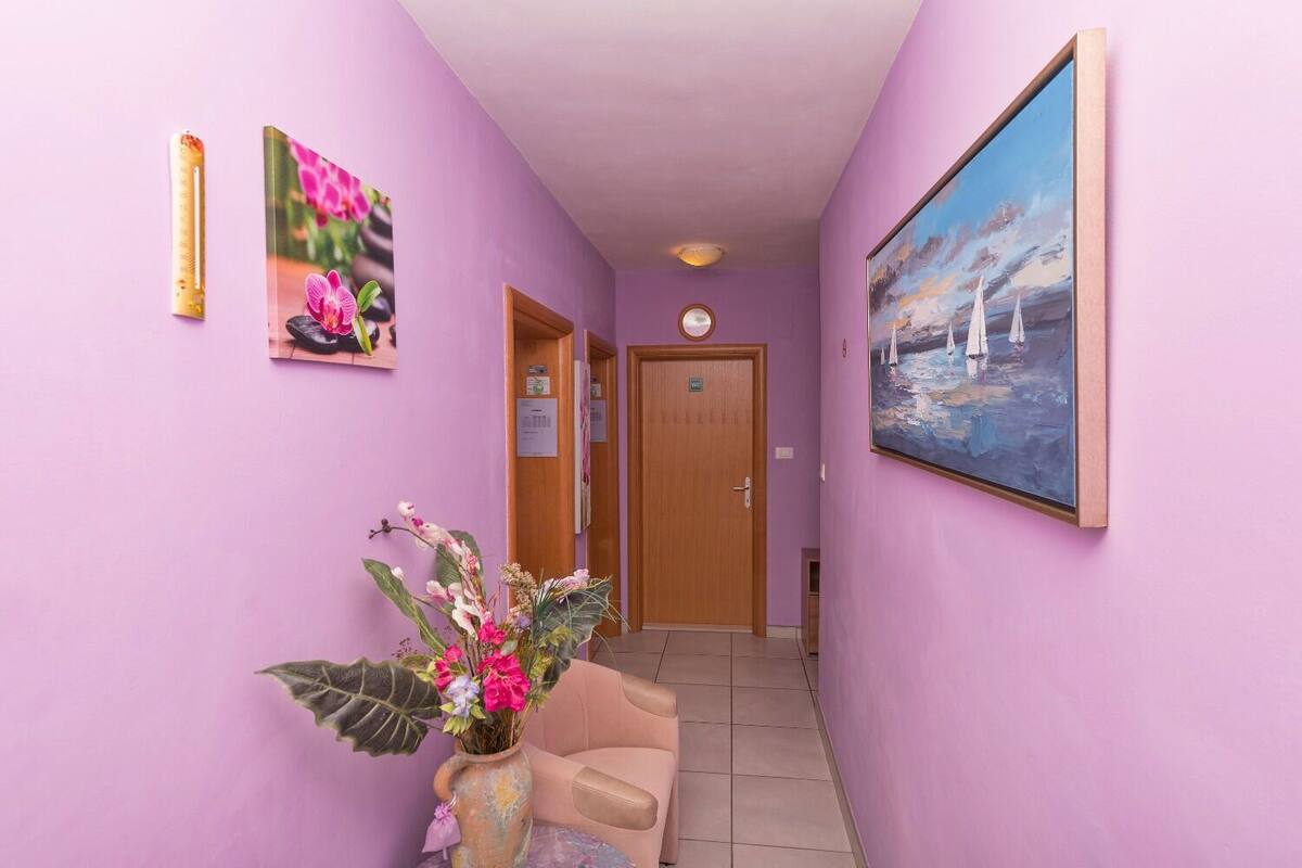 Un couloir avec un mur violet, une porte en bois, un canapé beige, un vase rempli de fleurs et un tableau représentant des voiliers.