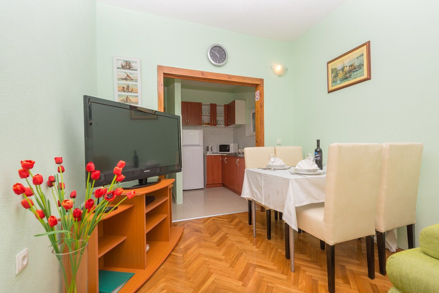 Un ambiente compatto con una TV su un mobile in legno, un tavolo da pranzo con sedie, una zona cucina e una porta che conduce in un'altra stanza.