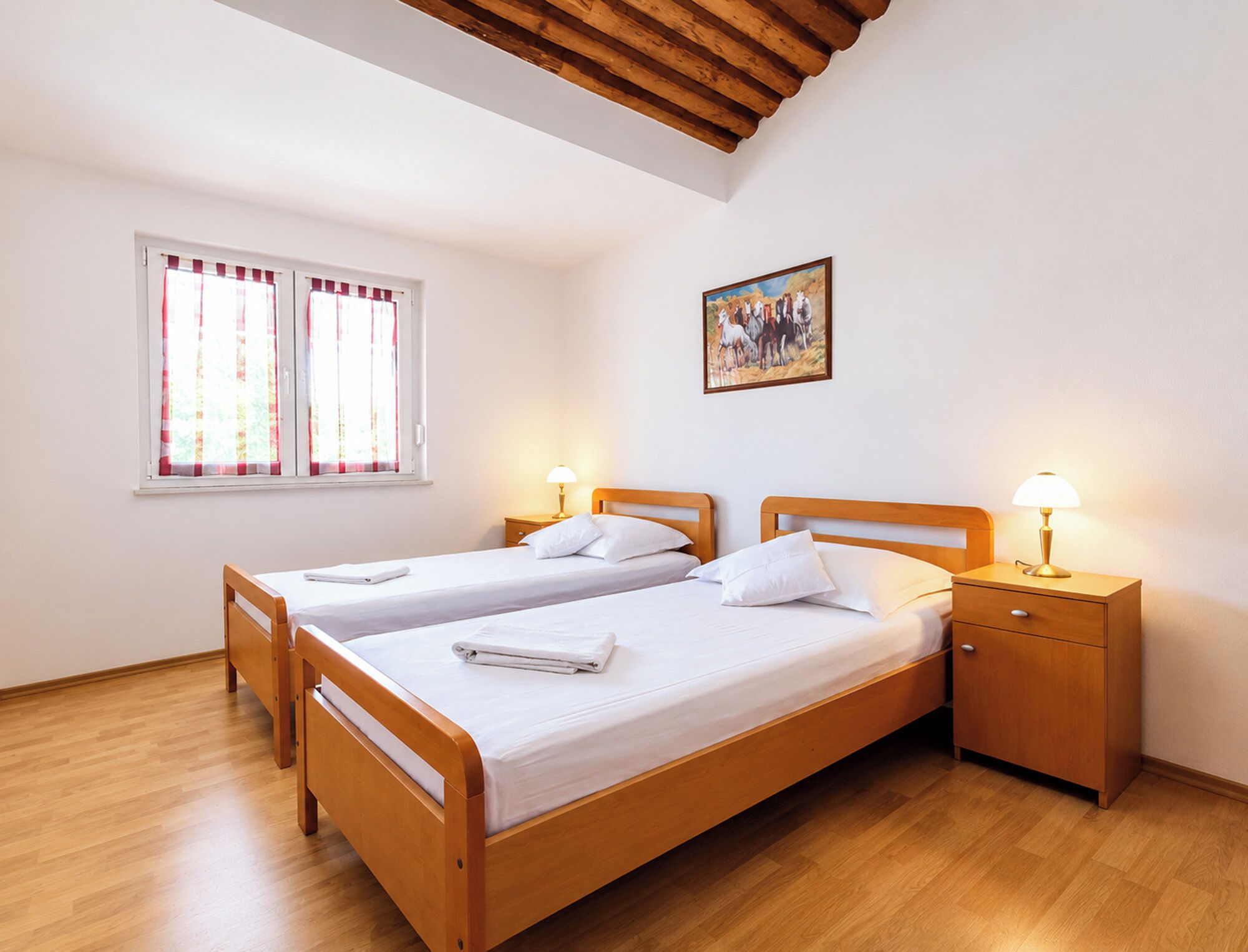 A double bed with white linens, two bedside tables with lamps, a window with curtains, and a painting on the wall.