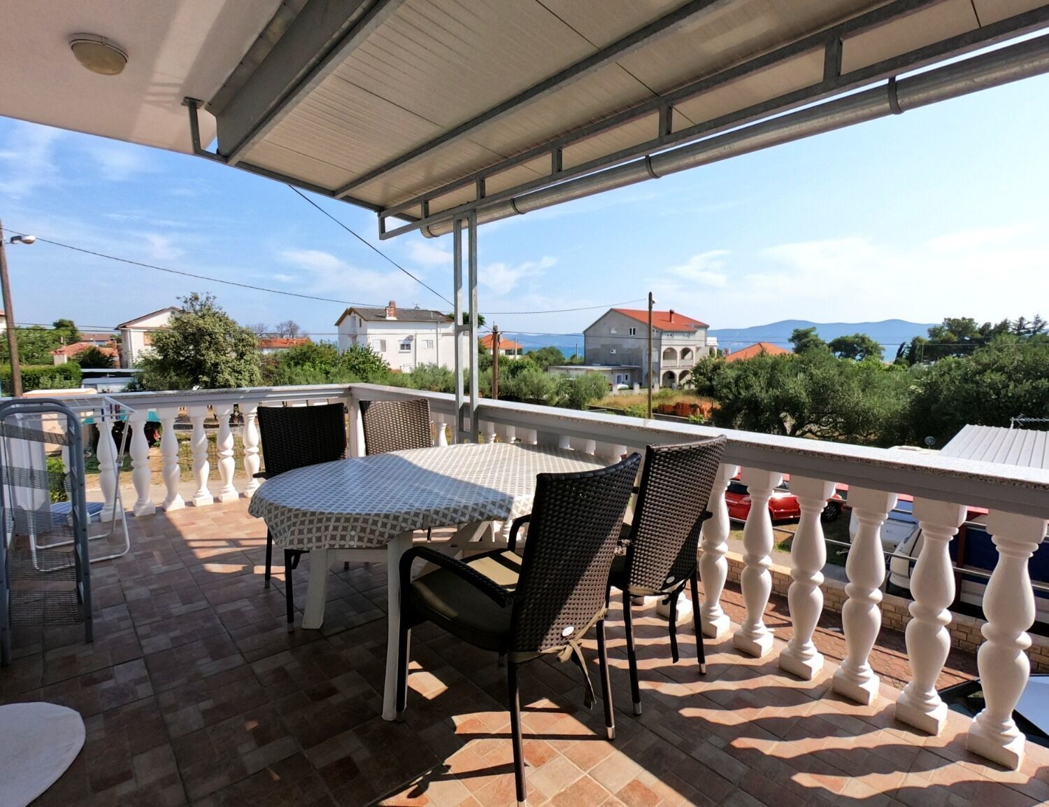 A covered outdoor patio with a table and chairs, a white balustrade, and a view of houses and mountains in the background.