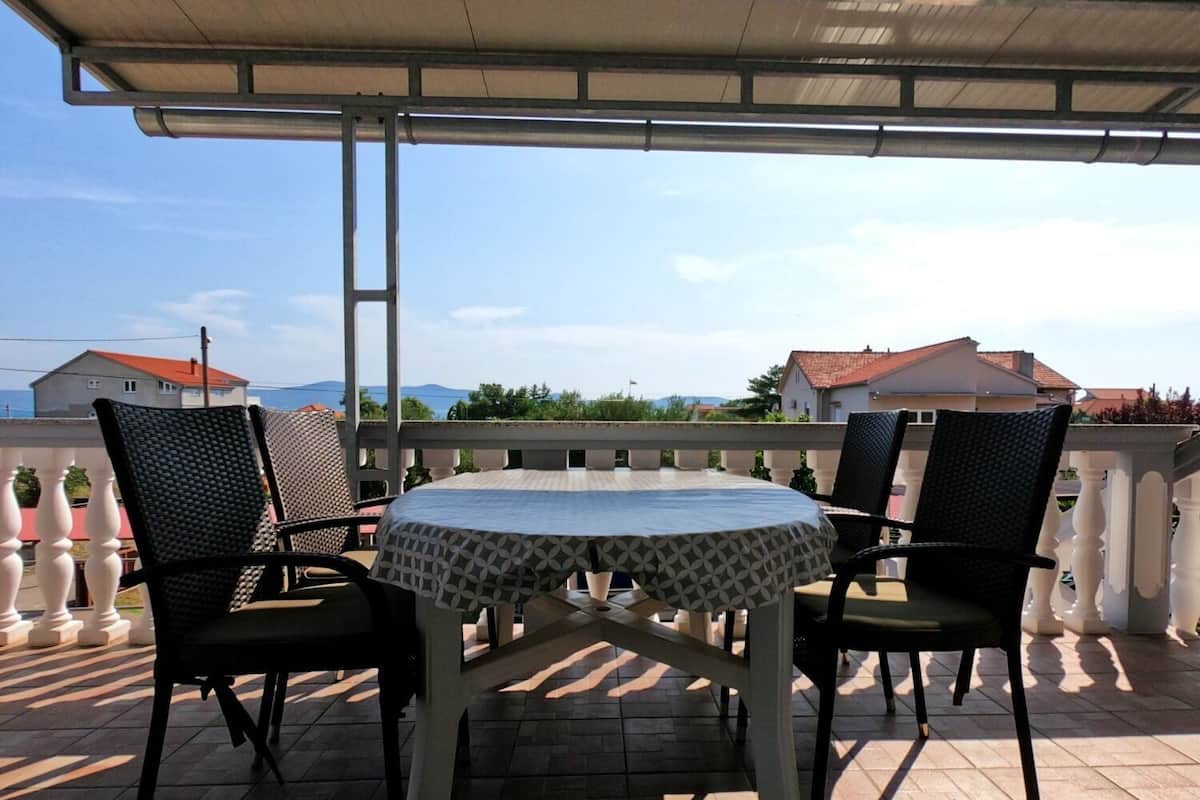 Un patio techado al aire libre con una mesa redonda y cuatro sillas, con vistas a una zona residencial con casas y montañas al fondo.