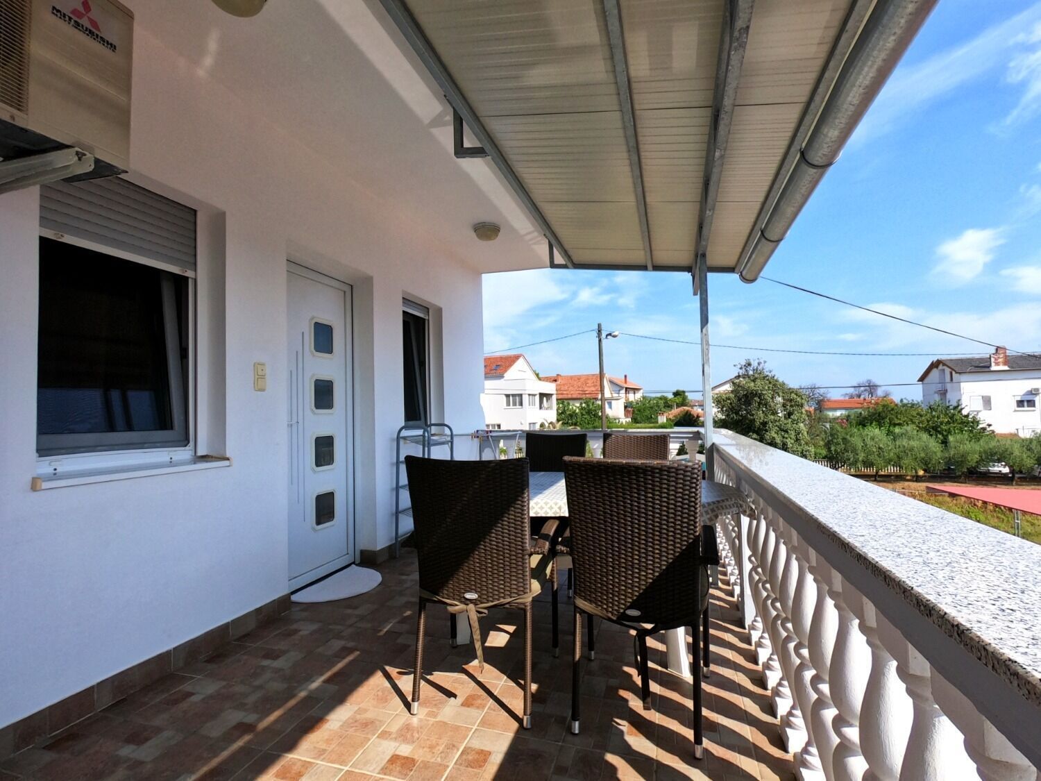 A covered balcony with a table and chairs, a door, and a view of neighboring houses.