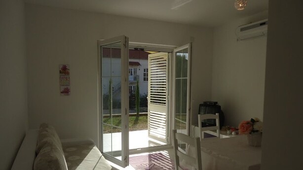 A room with a white sofa, a dining table with a chair, and a sliding glass door leading to an outdoor area. - Apartments Mirjana (Pakostane)