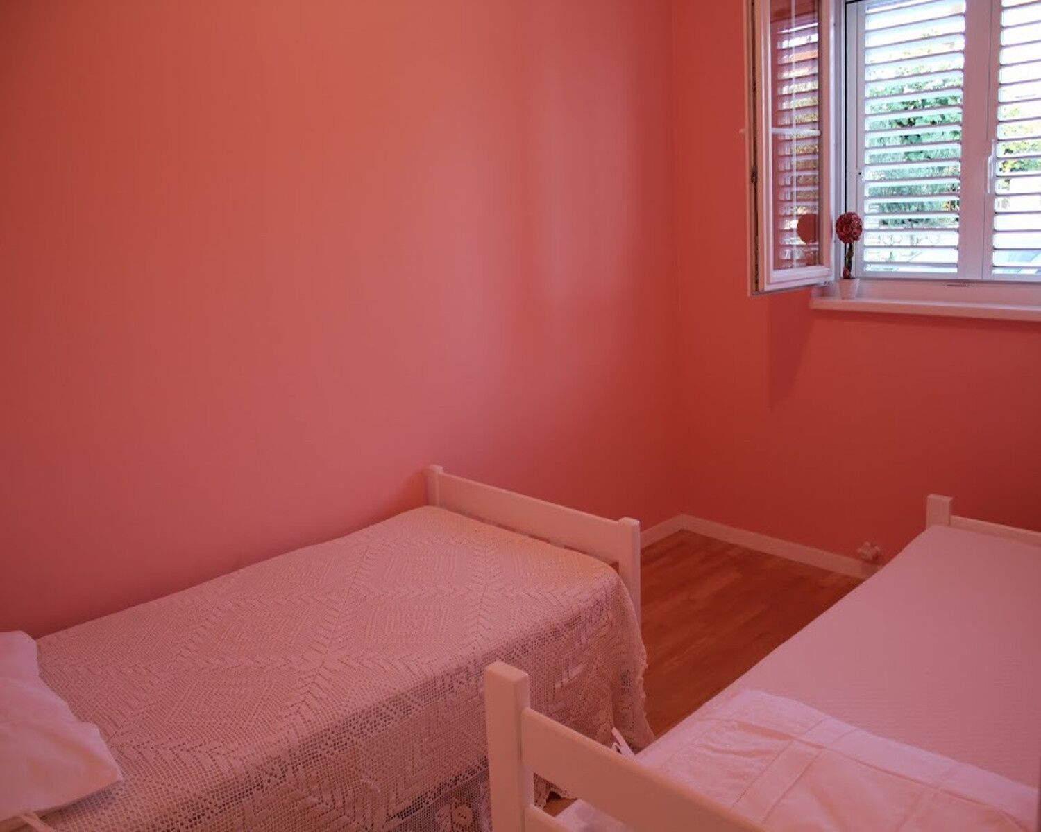 Una habitación pequeña con paredes rosas, dos camas individuales, una ventana con persianas y una mesita con un florero.