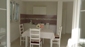 A dining area with a table and four chairs, a kitchen in the background, and a window with blinds. - Apartments Mirjana (Pakostane)