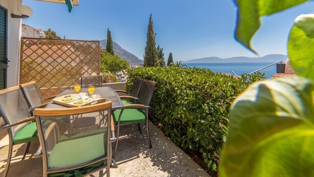 An outdoor patio with a table set for two, surrounded by greenery and overlooking a scenic view of the sea and mountains. - Apartments Horizont (Gradac)