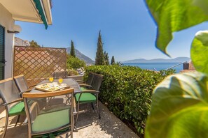 An outdoor patio with a table set for two, surrounded by greenery and overlooking a scenic view of the sea and mountains.