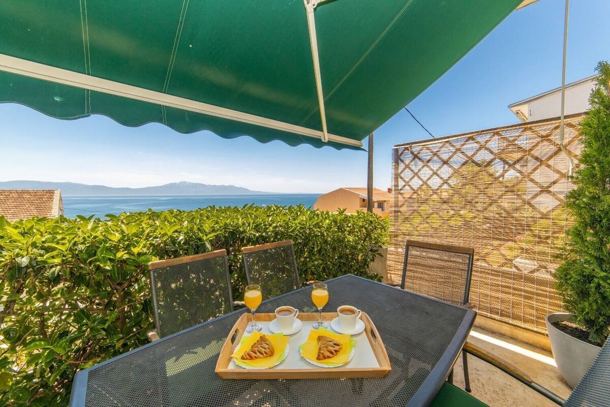 A terrace with a table set for breakfast, including pastries, coffee, and juice, overlooking a scenic view of the sea and mountains.