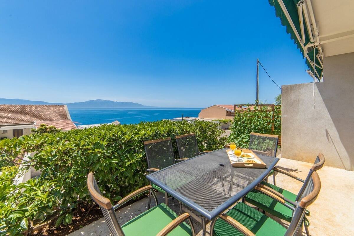 A balcony with a table and chairs, overlooking a coastal view with buildings and the sea.
