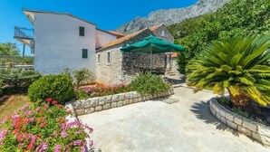 A two-story building with a stone facade and a white upper section. There is a green umbrella over a seating area. - Apartments Horizont (Gradac)
