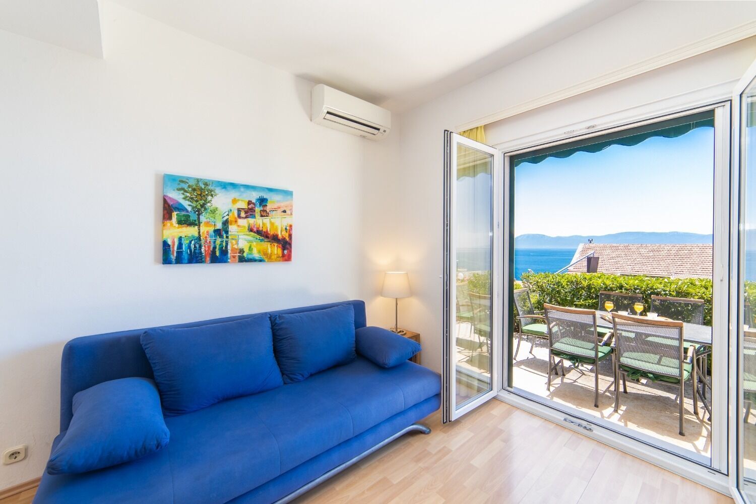 A room with a blue sofa, a painting, and a view of the sea through a glass door.