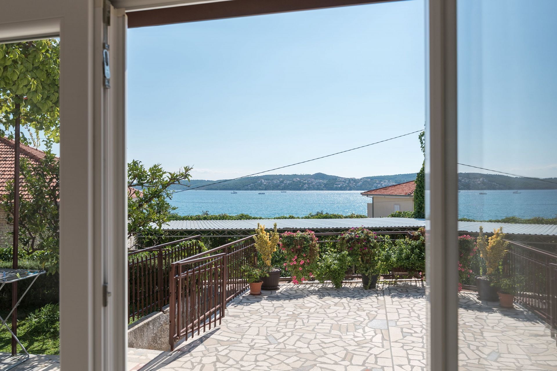 Blick von einem Balkon auf ein Gewässer mit Booten, einen fernen Berg und eine mit Töpfen bepflanzte, mit Fliesen ausgelegte Terrasse.