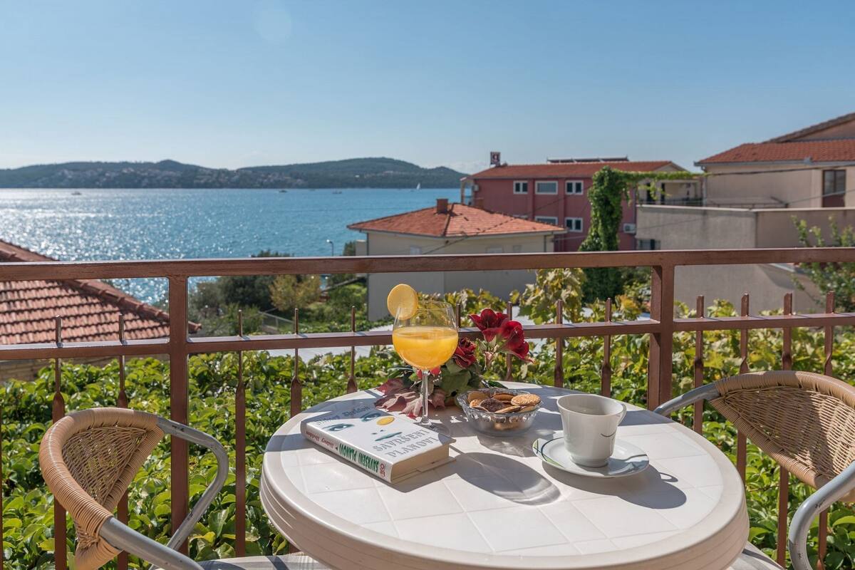 A balcony with a table set for two, featuring a book, a glass of orange juice, a cup, and a bowl of cookies.