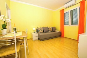 A small, brightly colored room with a wooden floor, a sofa, a table with a bottle and glasses, and a window with orange curtains. - Apartment Neno (Preko)