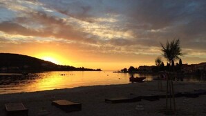 A beach with a sunset over water, a few trees, and a distant shoreline with buildings. - Apartment Neno (Preko)