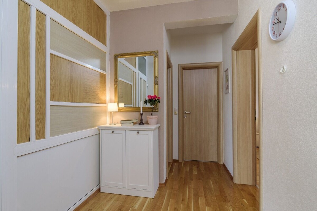 A narrow hallway with wooden flooring, a white cabinet with a mirror, a wall clock, and a door at the end.