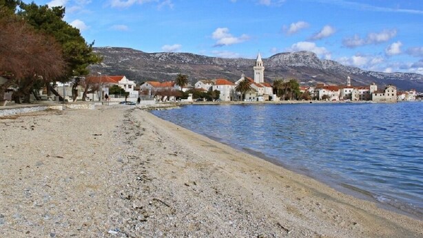 Un pueblo costero con playa de guijarros, edificios con tejados rojos y una iglesia con campanario.