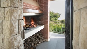 Ein Kamin aus Stein und Ziegel mit Feuer, befindet sich auf einem Balkon mit Metallgeländer und Grünpflanzen im Hintergrund.