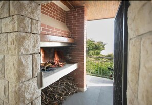 Ein Kamin aus Stein und Ziegel mit Feuer, befindet sich auf einem Balkon mit Metallgeländer und Grünpflanzen im Hintergrund.